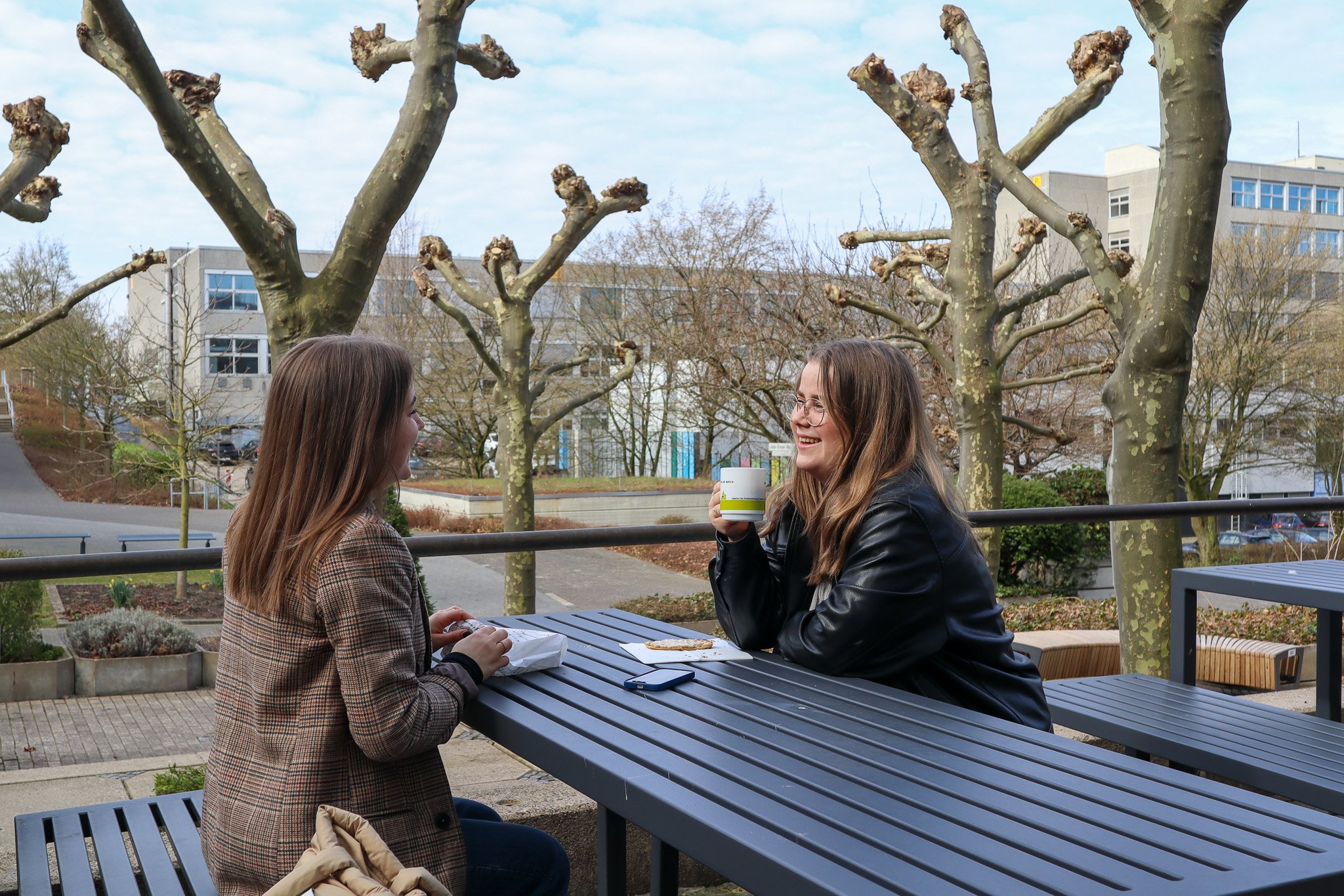 Zwei Personen sitzen an einem Tisch im Freien, umgeben von beschneiden Bäumen, und unterhalten sich bei Snacks und Getränken.