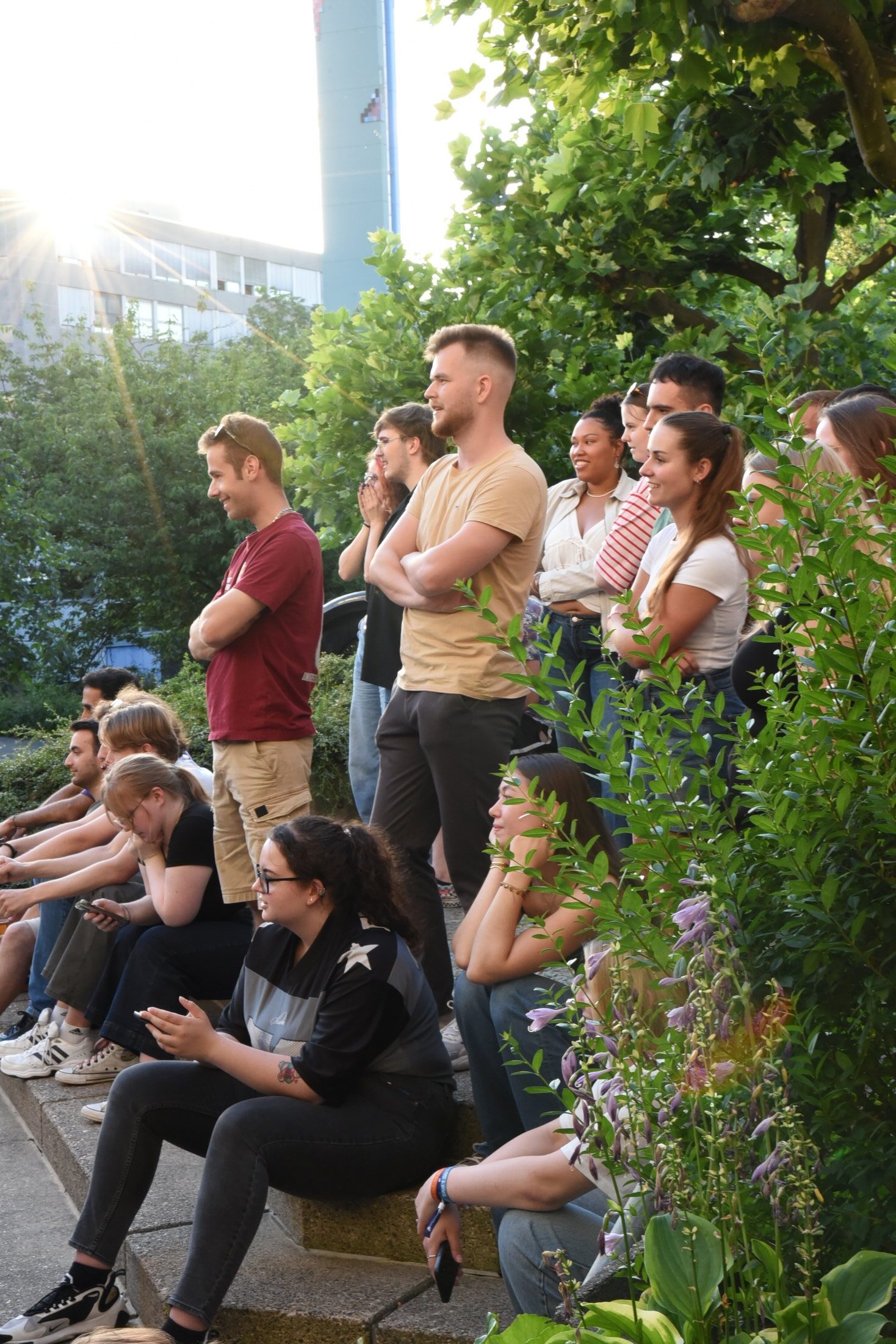 A group of young people sit and stand in a green outdoor area, surrounded by plants and light.