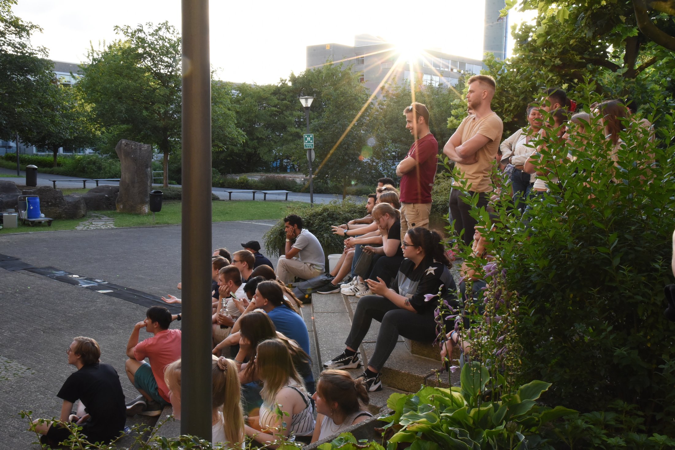Gruppenvon Menschen sitzt und steht in einem begrünten Außenbereich, teilweise vor einer Leinwand, bei Sonnenuntergang.