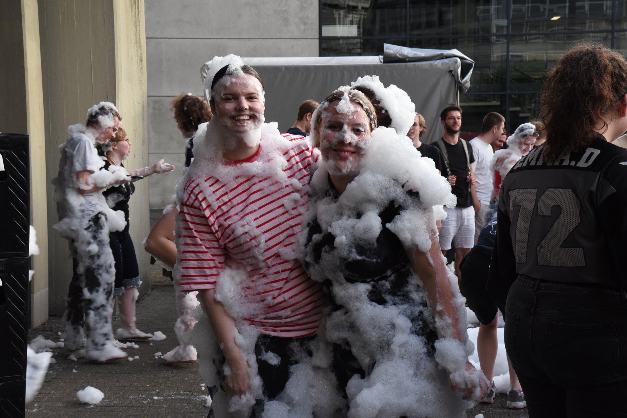 Zwei Personen stehen in Schaum, umgeben von weiteren Menschen. Der Boden ist mit Schaum bedeckt.