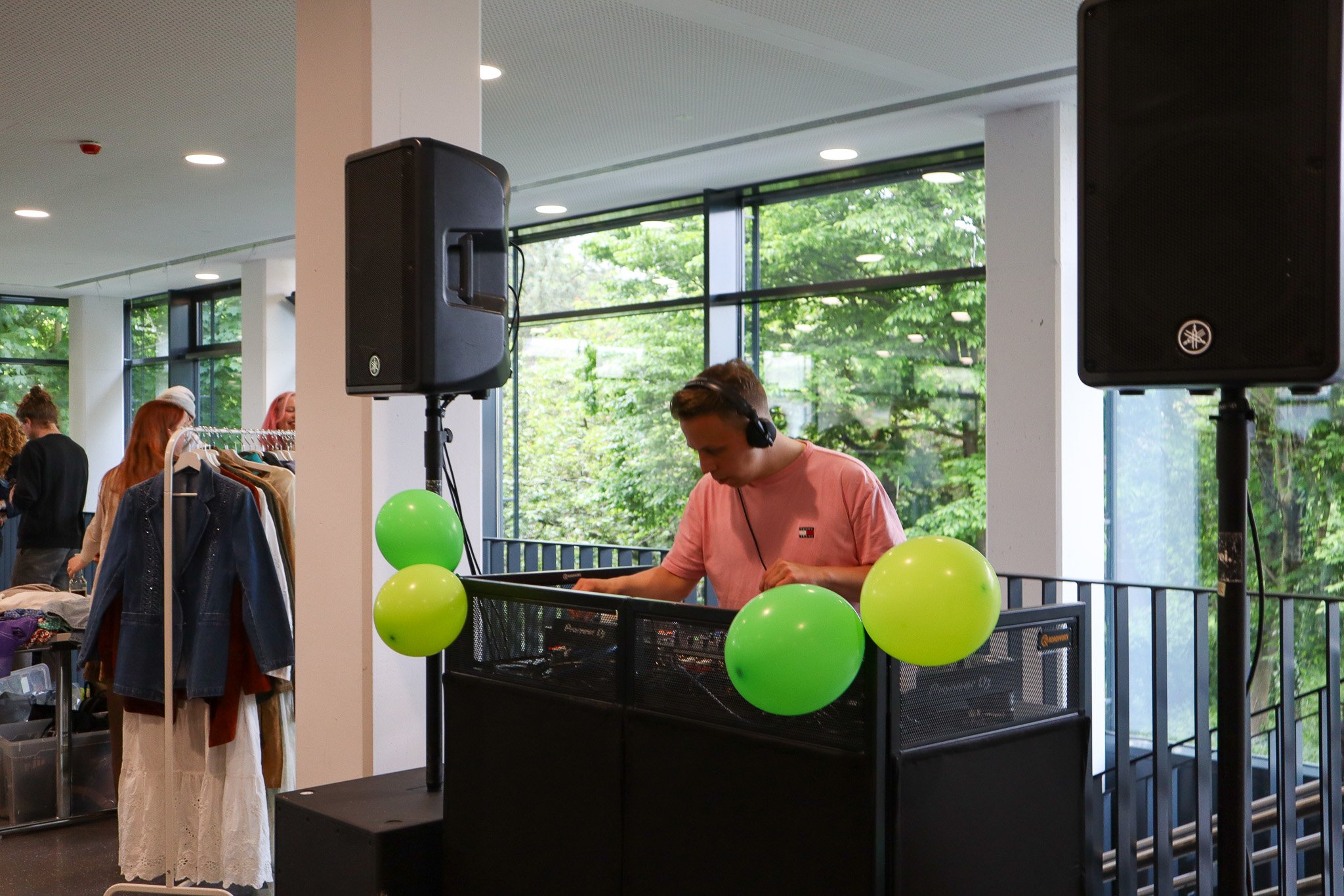 Ein DJ mit Kopfhörern spielt Musik an einem Mischpult, umgeben von grünen Luftballons und einem sonnigen Raum.