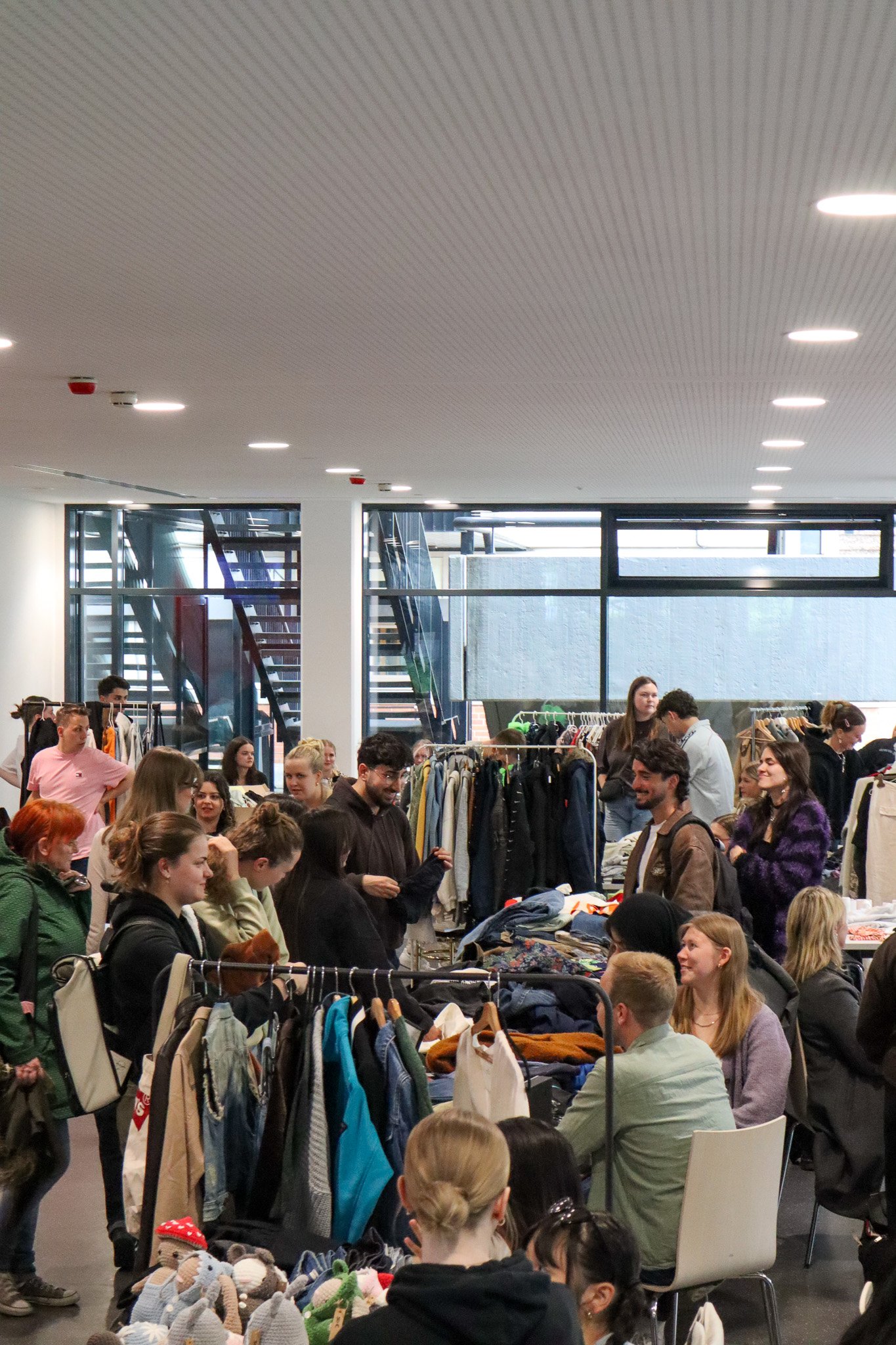 In einem Raum stehen Menschen um Ständer mit Kleidung. Einige unterhalten sich, andere schauen in die Regale.