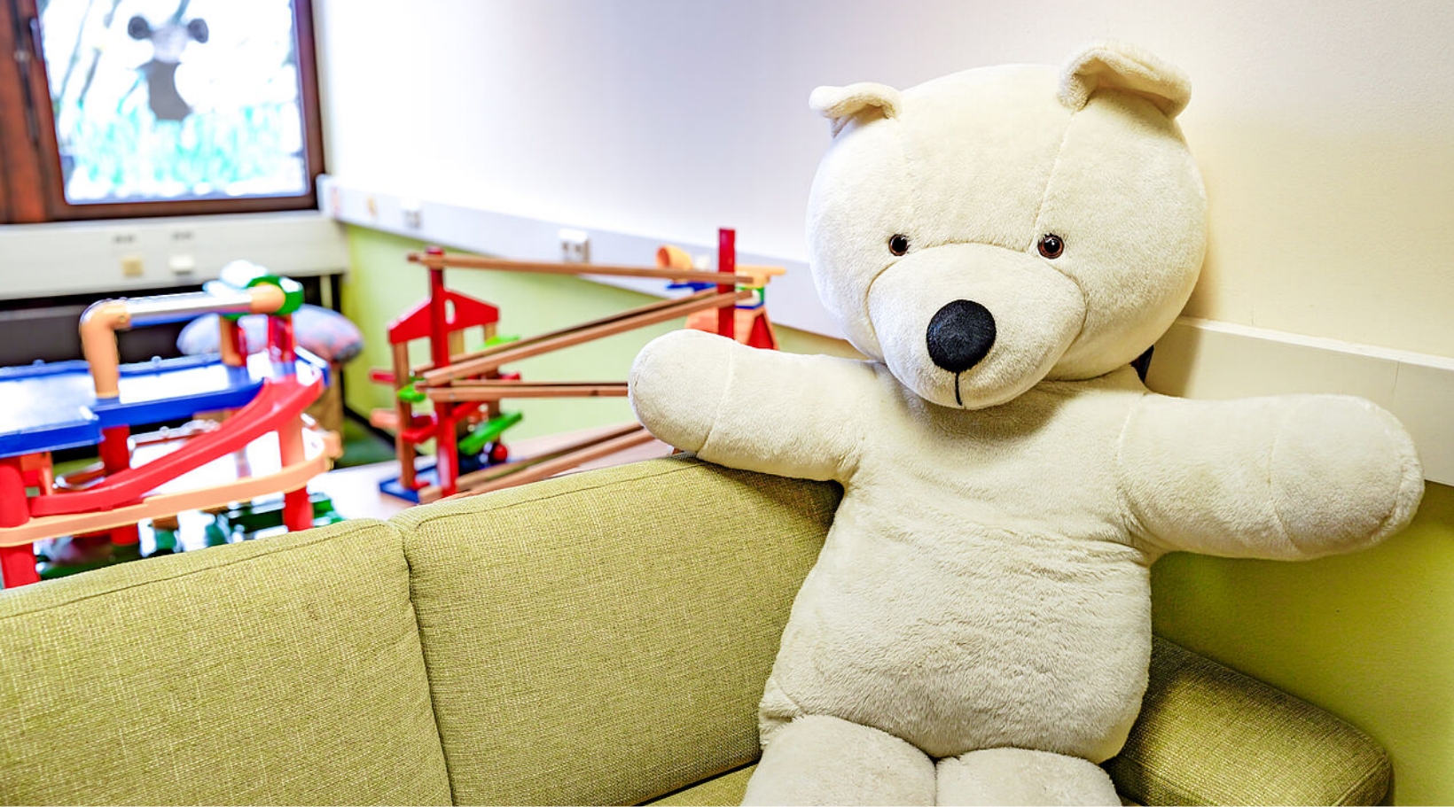 A large white teddy bear can be seen in the foreground, sitting on a green sofa. There are various toys in the background.