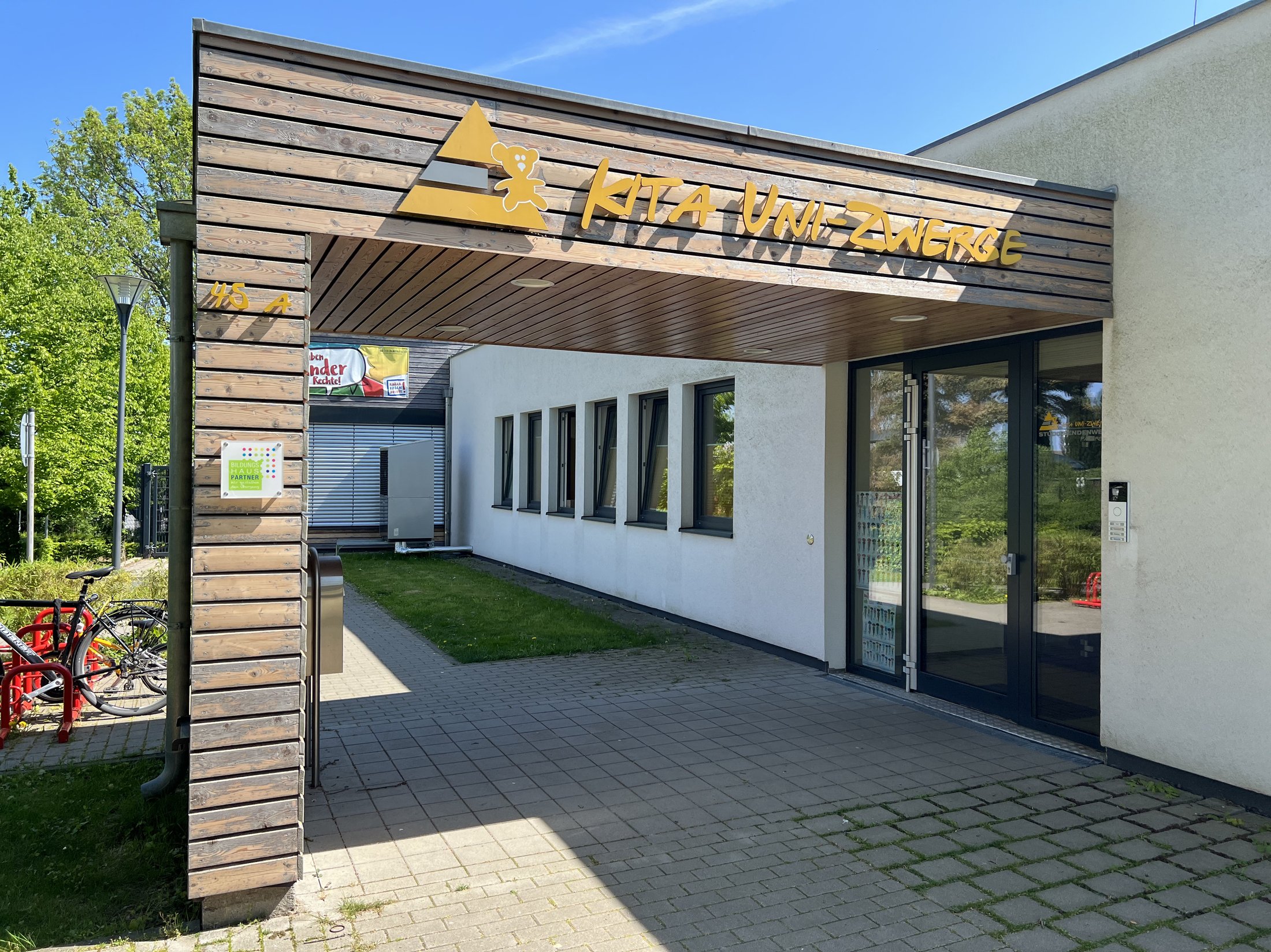 Entrance to the Uni-Zwerge daycare centre with wooden roof, large glass door and seating area, surrounded by greenery and bicycles.