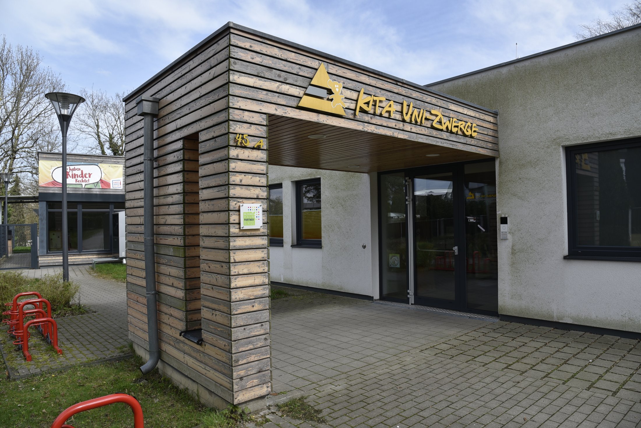Entrance to the daycare centre with wooden panelling and a large sign, next to it a terrace and bicycle racks. Bright, cloudy sky.