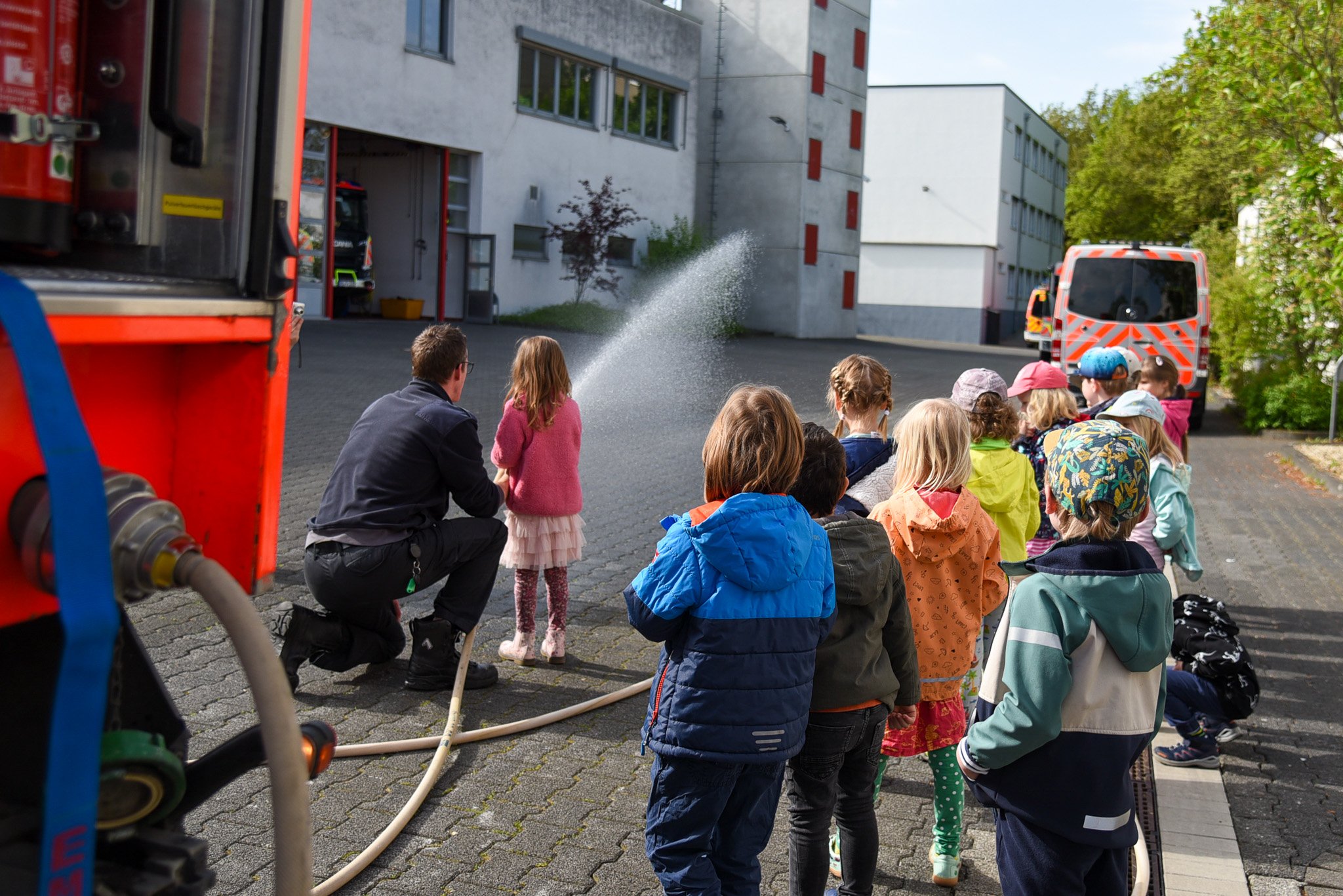 Children stand in a row while a fireman shows a small group how to spray with a fire hose.