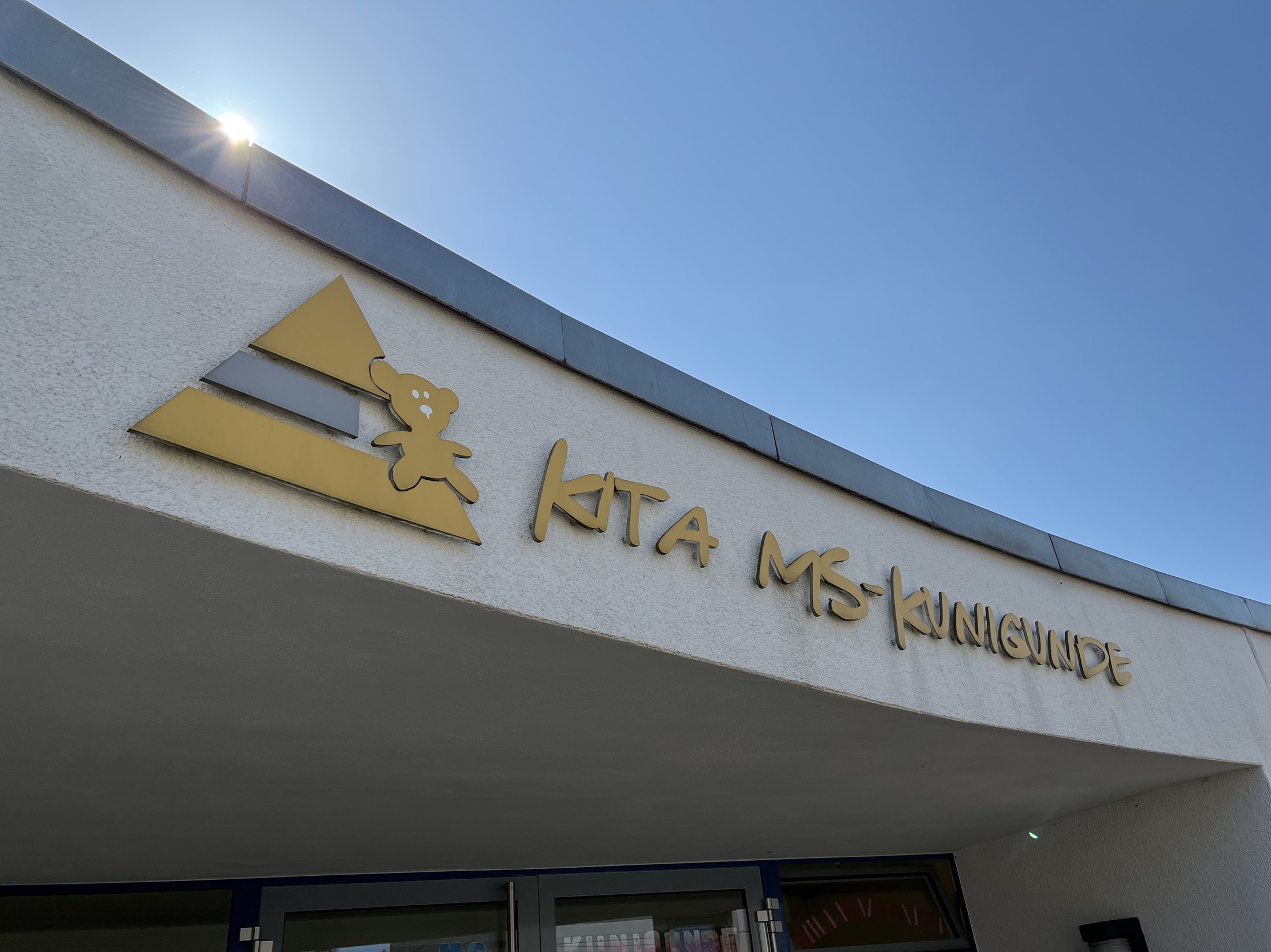 Sign for the MS-Kunigunde daycare centre with a bear motif and colourful geometric shapes against a blue sky.