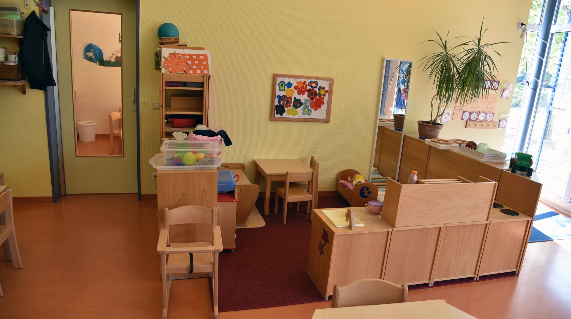 Bright group room with tables, chairs, toys and a plant. A work of art hangs on the wall.