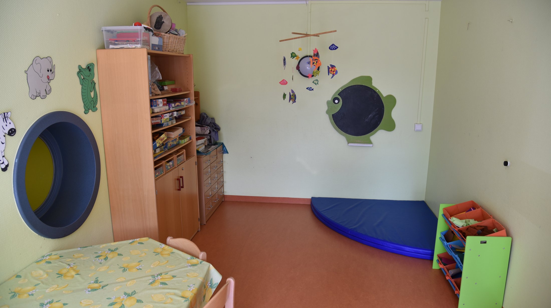 Playroom with colourful walls, a round window, toys on shelves and a blue gym mat.