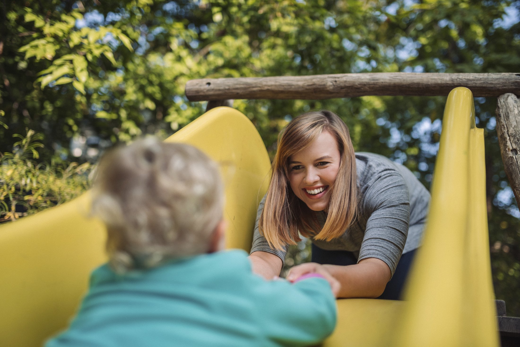 A person sits on a slide and shakes hands with a child. Trees surround the play area.