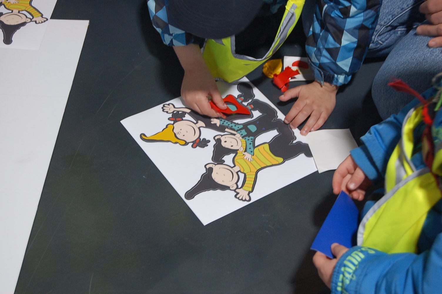 Children place coloured shapes on a picture of three cheerful characters with different hats.