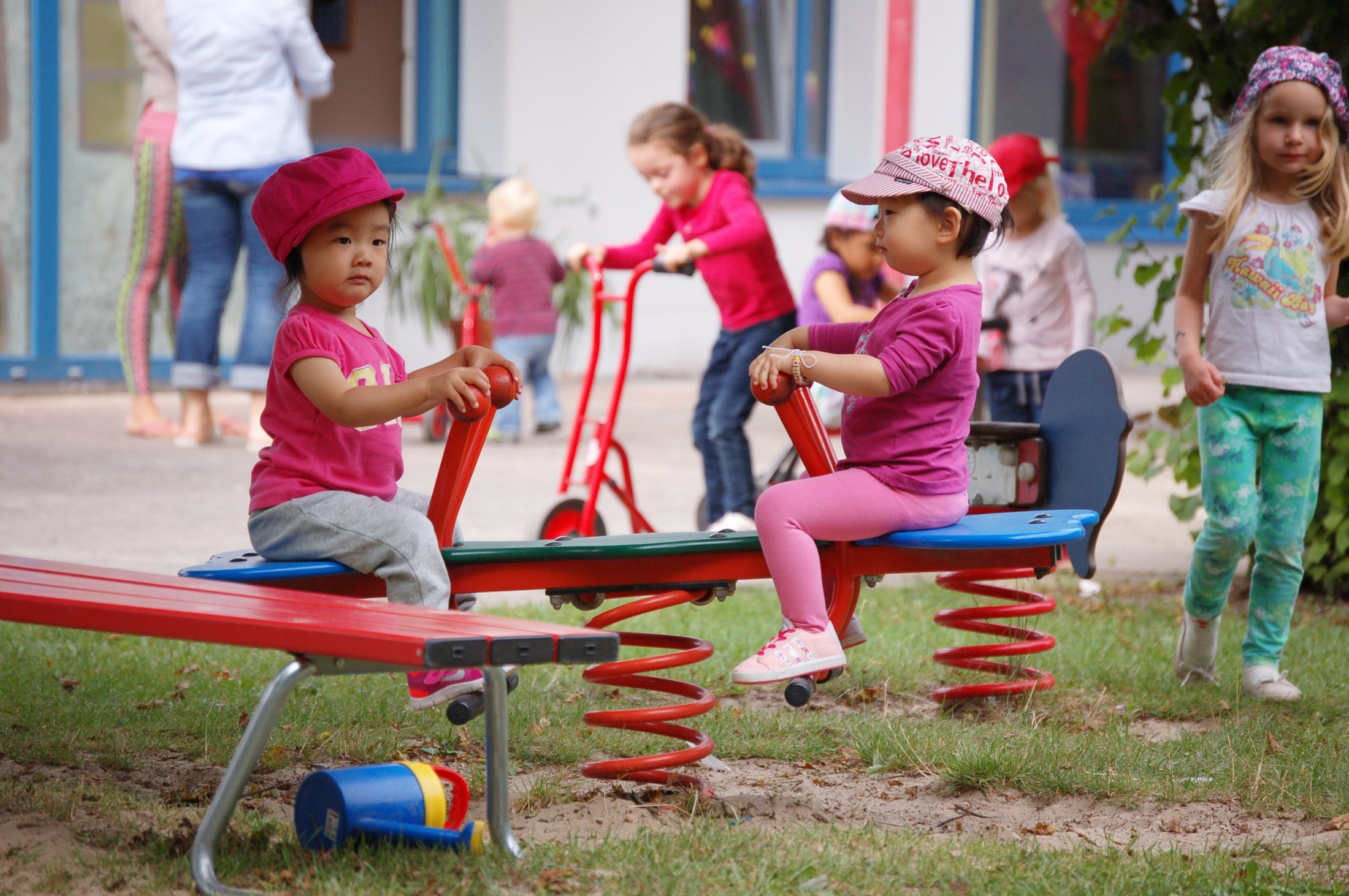 Two children on a seesaw outdoors, surrounded by other children playing and a toy in bright colours.