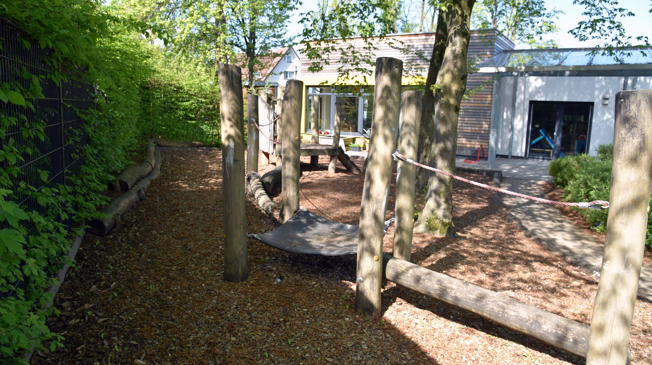 Playground with wooden frames, a balancing area and trees. In the background is a building with large windows.