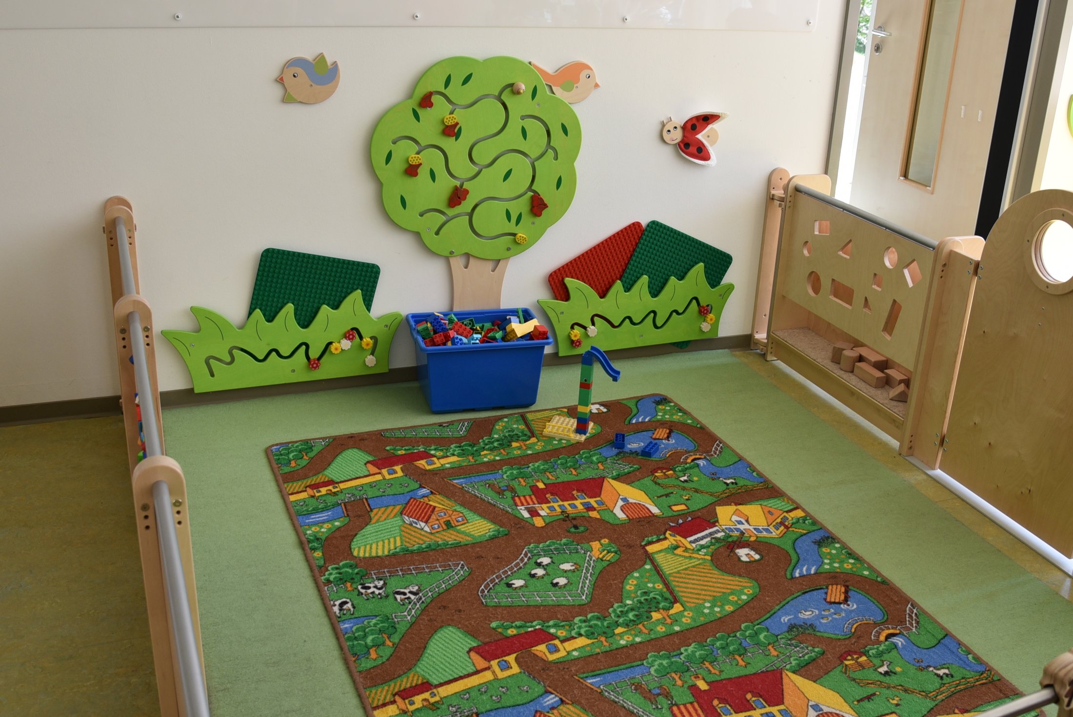 A colourful play area with carpet, toy box, wall play and green tree design.