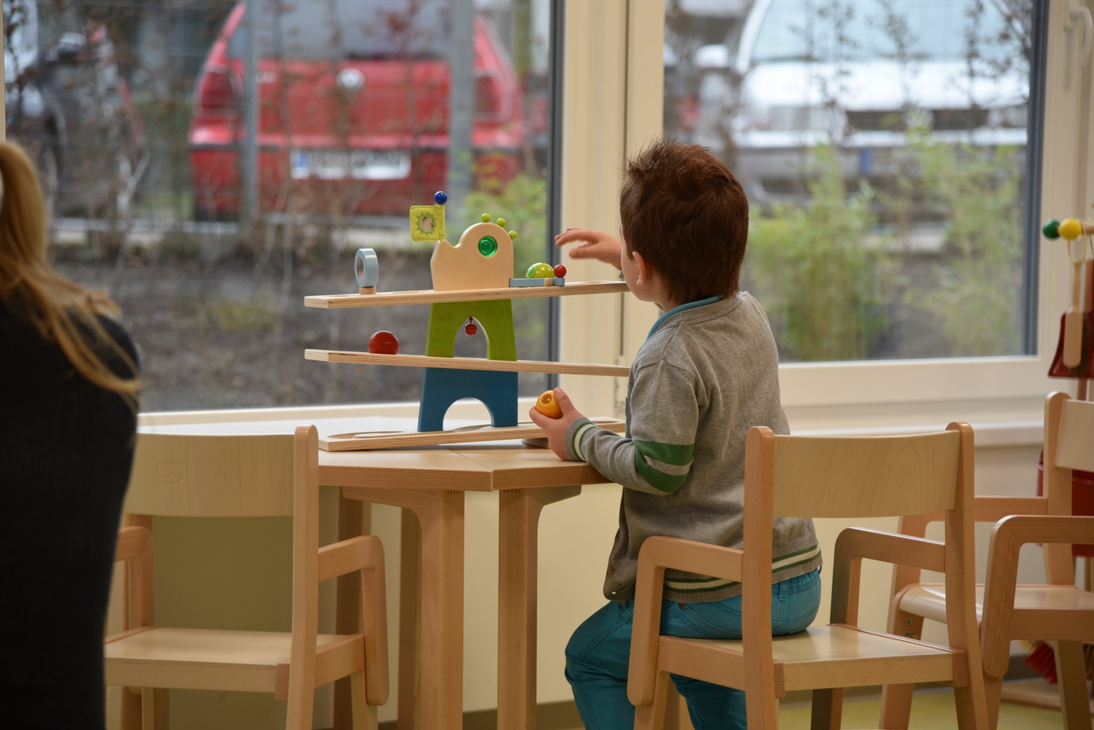 A child plays at a table with a colourful wooden toy that has several levels. Background with windows and cars.