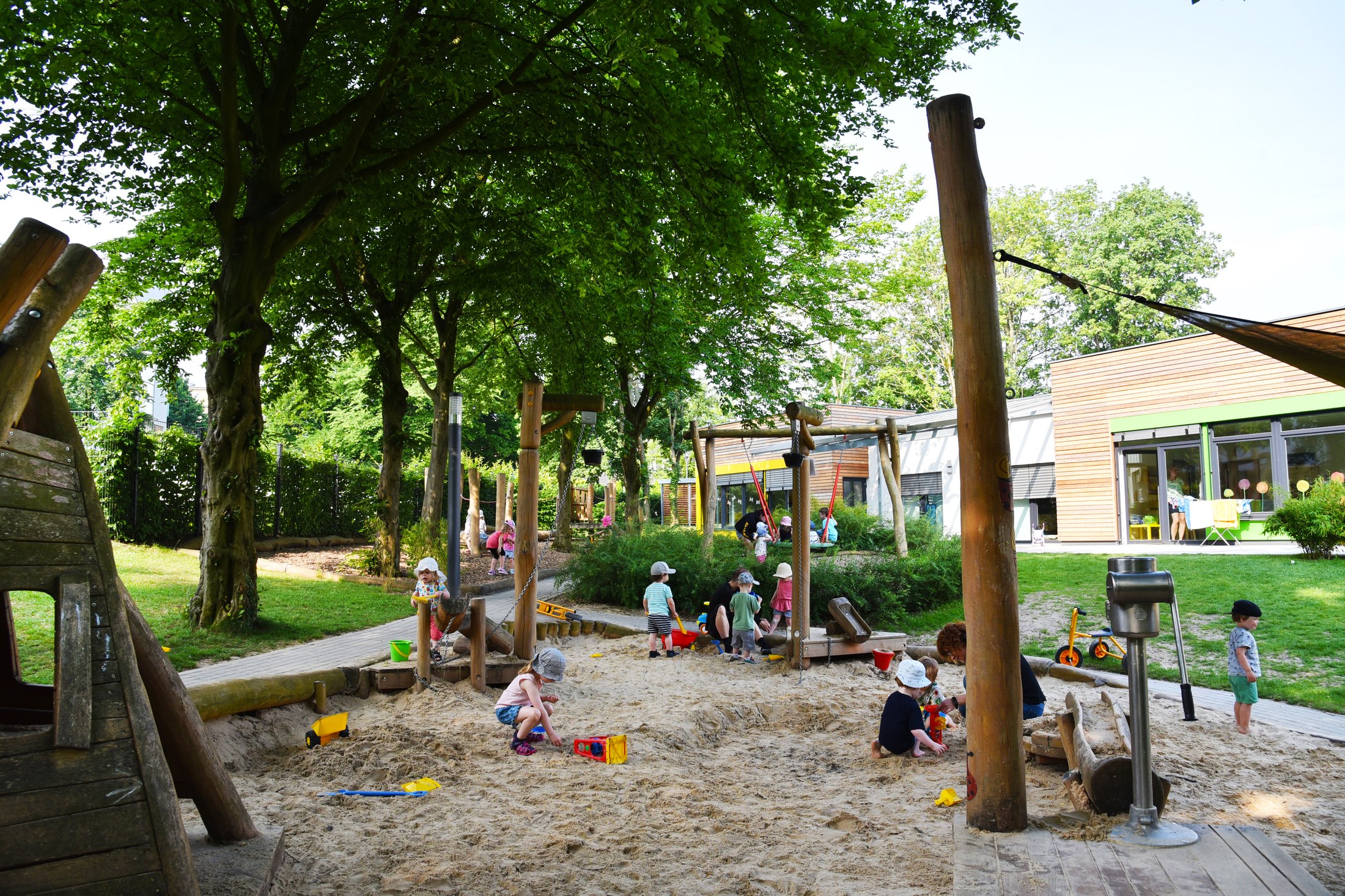 Playground with sandpit, play equipment and children playing outdoors. Trees and a building in the background.