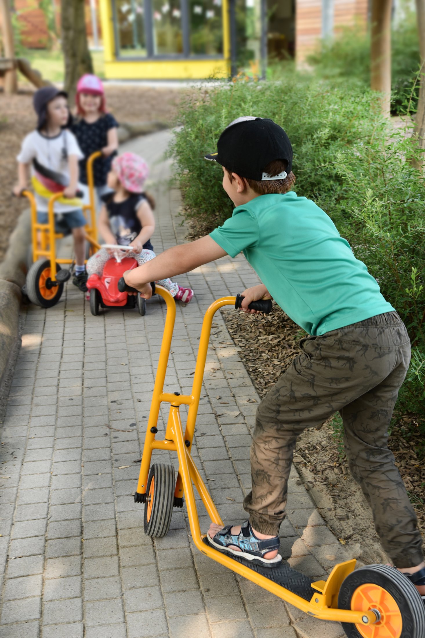 Children ride on a paved path with various vehicles, surrounded by green areas and a building in the background.
