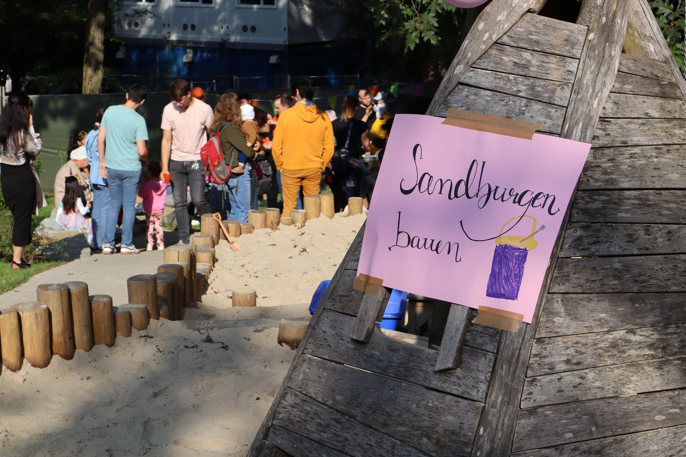 A wooden play equipment in the foreground with a colourful sign that reads "Building sandcastles". People and a sand area in the background.