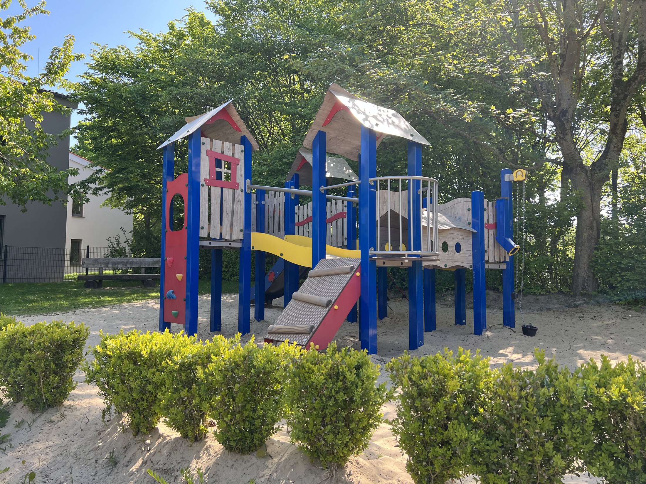 Play elements with slides and climbing facilities in a sandy play area surrounded by bushes and trees.
