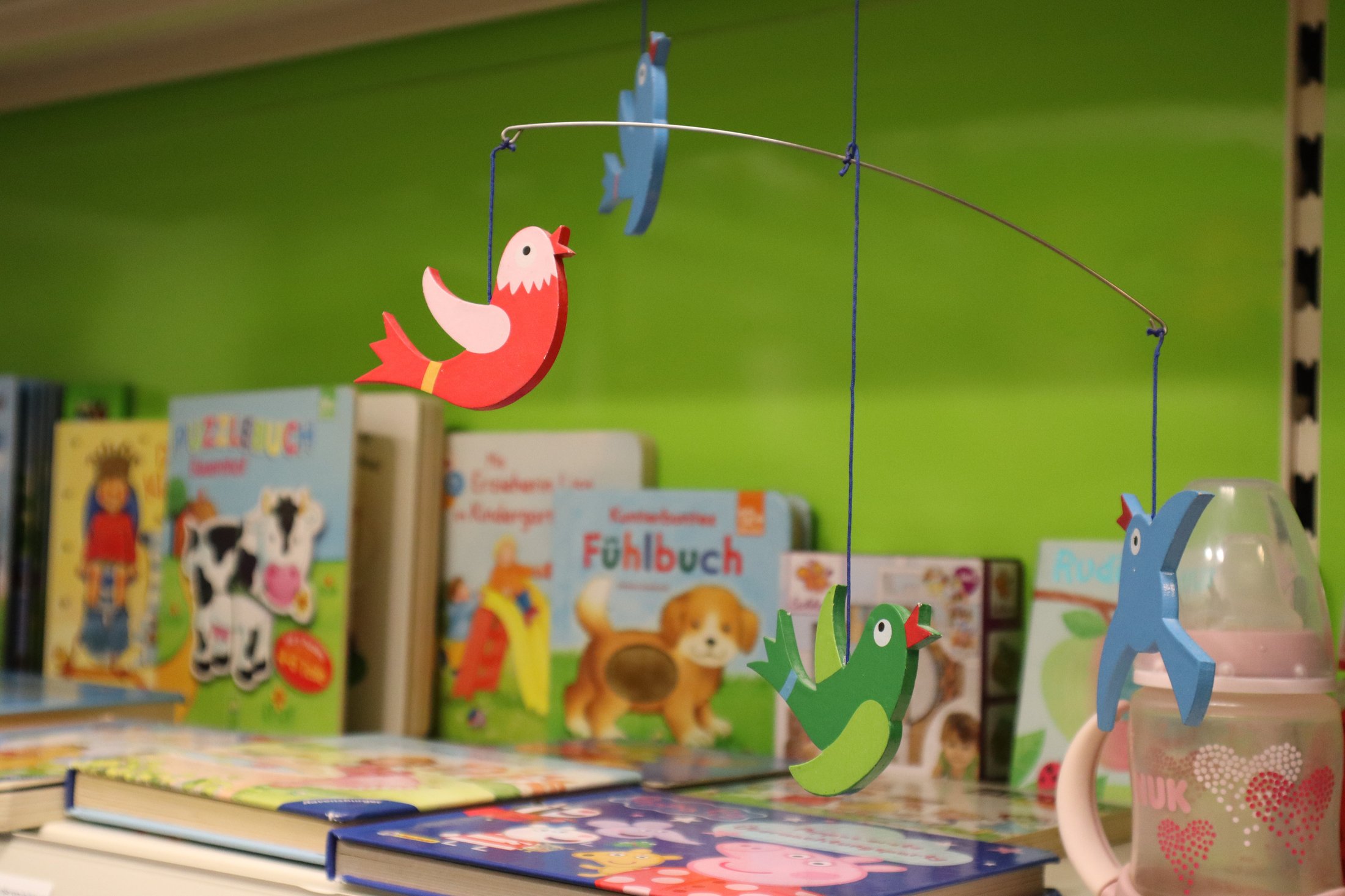 Buntes Mobile mit Vögeln hängt über einer Vielzahl von Kinderbüchern auf einem Regal vor einer grünen Wand.