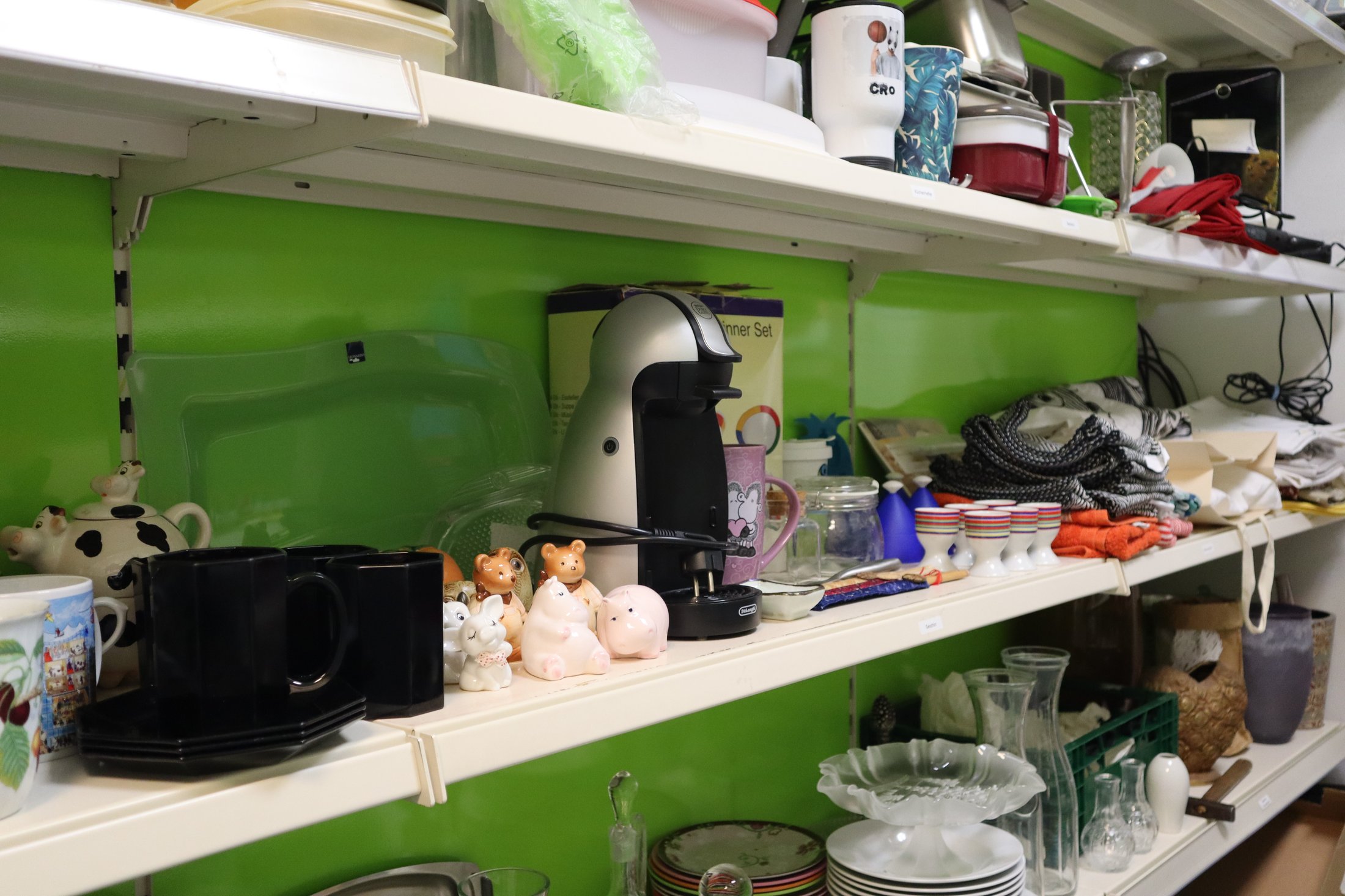 Ein Regal mit Haushaltswaren: Tassen, Geschirr, eine Kaffeemaschine und verschiedene Dekorationsartikel auf einem hellgrünen Hintergrund.