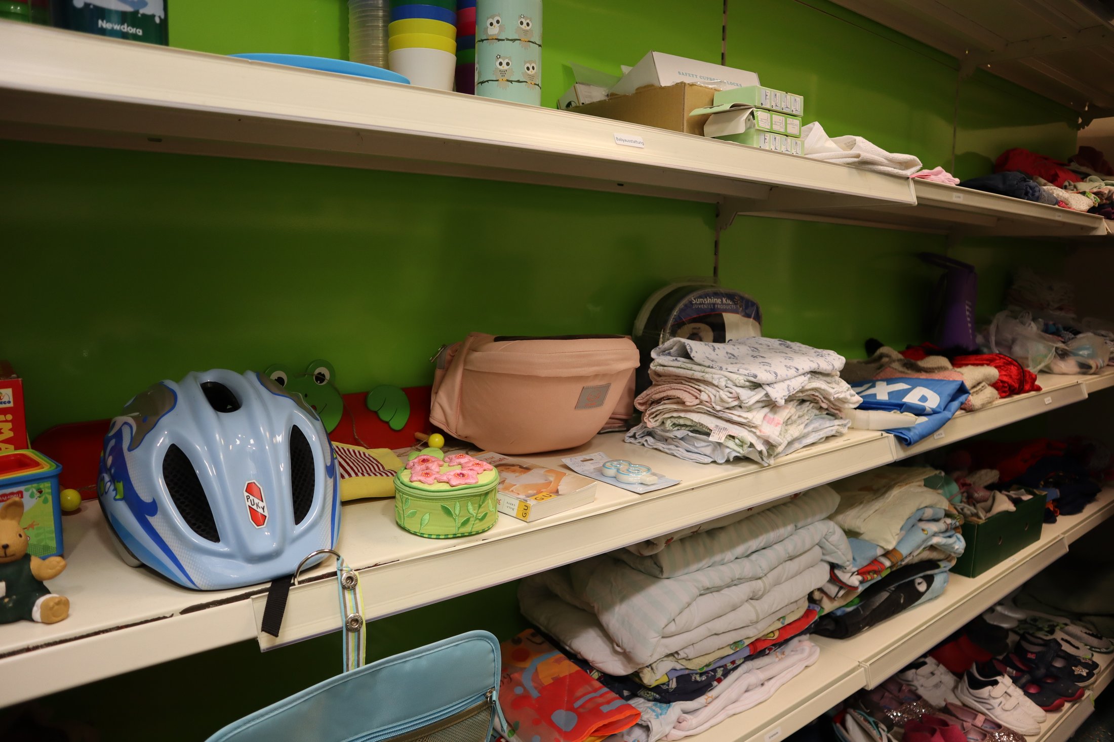 Regale mit Kleidung, Spielzeug und einem Fahrradhelm auf einem Regal mit grünem Hintergrund.