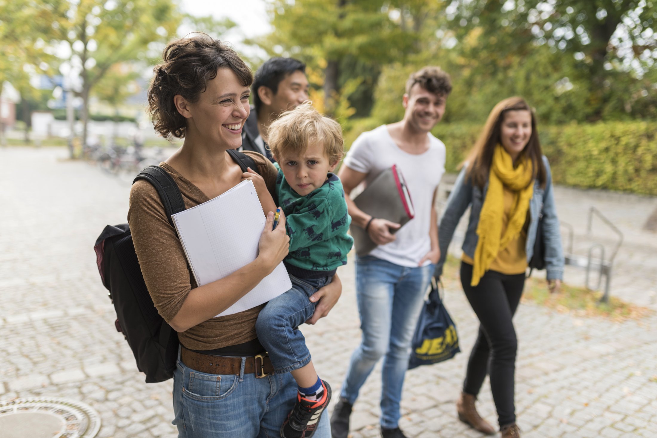 Group of four people on a paved path, one person carrying a child, others holding documents. Trees in the background.