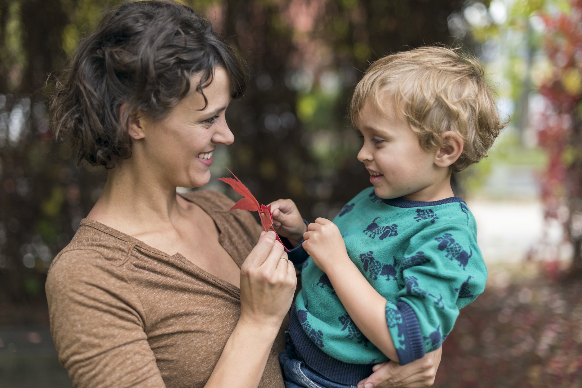 Eine Frau hält einen kleinen Jungen in den Armen, während sie ihm ein rotes Blatt zeigt. Bunte Blätter im Hintergrund.