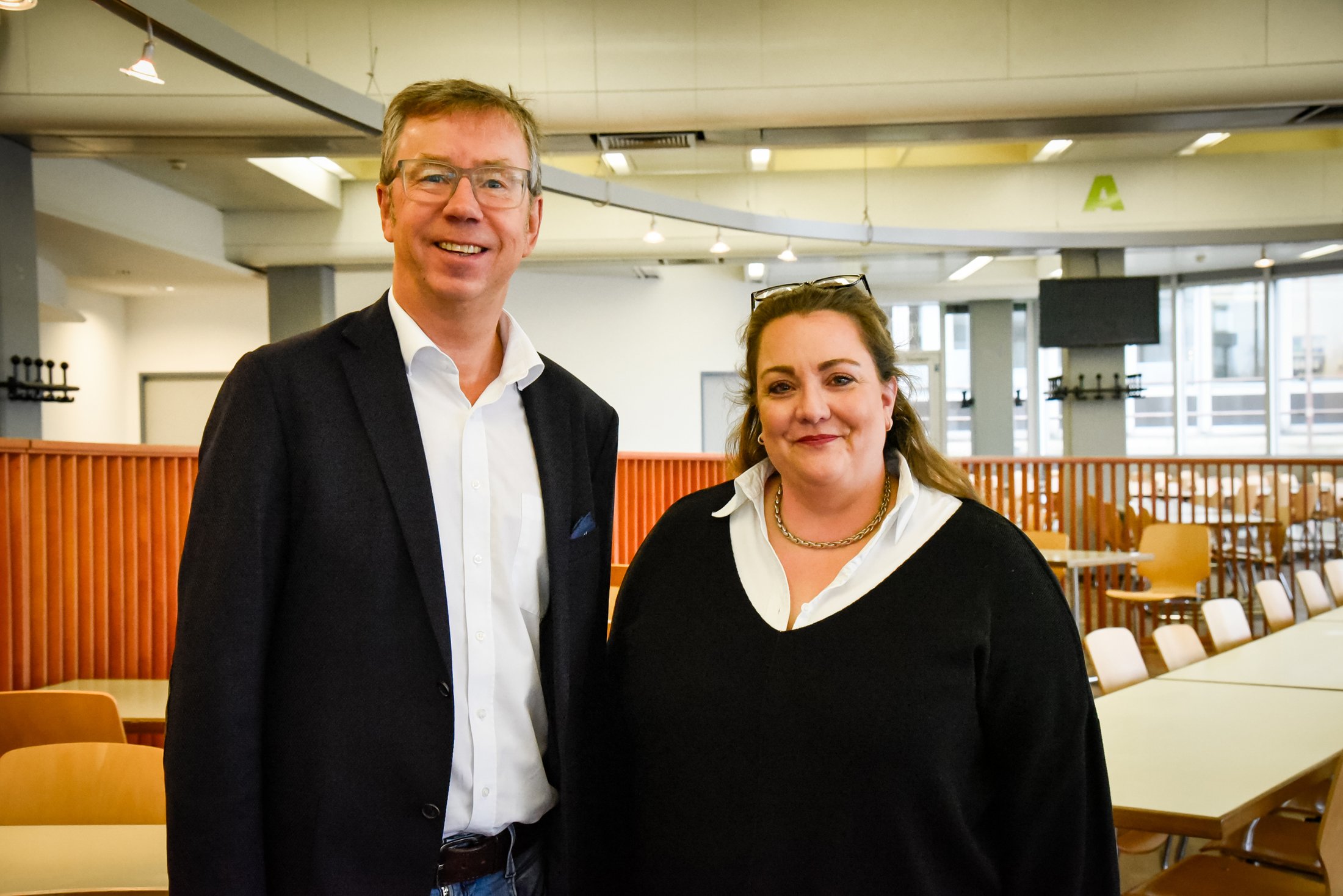 Two people smiling in a modern room with tables and chairs in the background. Bright, friendly atmosphere.